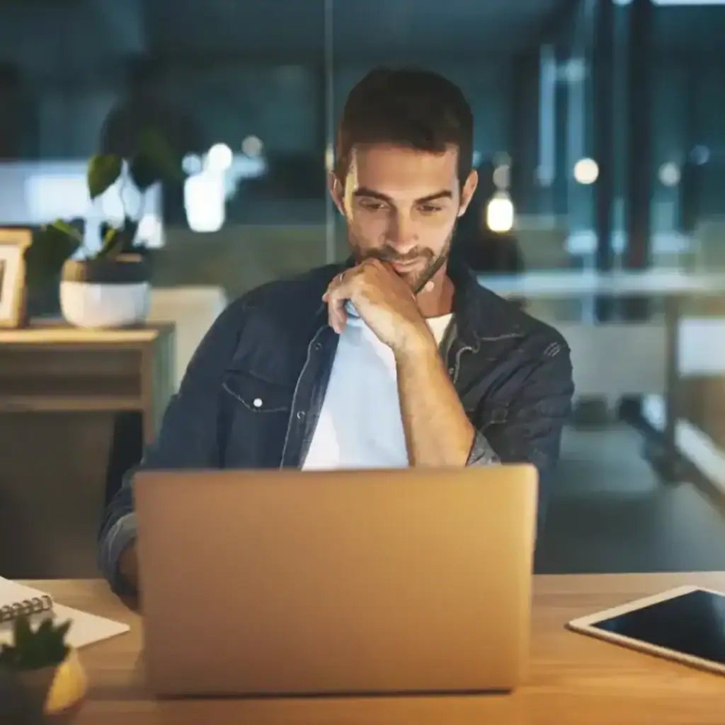 A man working on a laptop at a desk in a modern office at night.