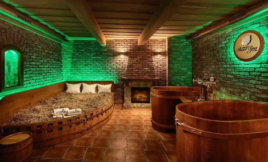 A cozy, dimly lit beer spa room with green ambient lighting. Two large wooden barrel bathtubs are on the right, and a straw-filled bed with white pillows and towels is on the left. A brick wall with a small fireplace is in the background, and a "Beer Spa" logo sign with a hop leaf is visible on the far right wall