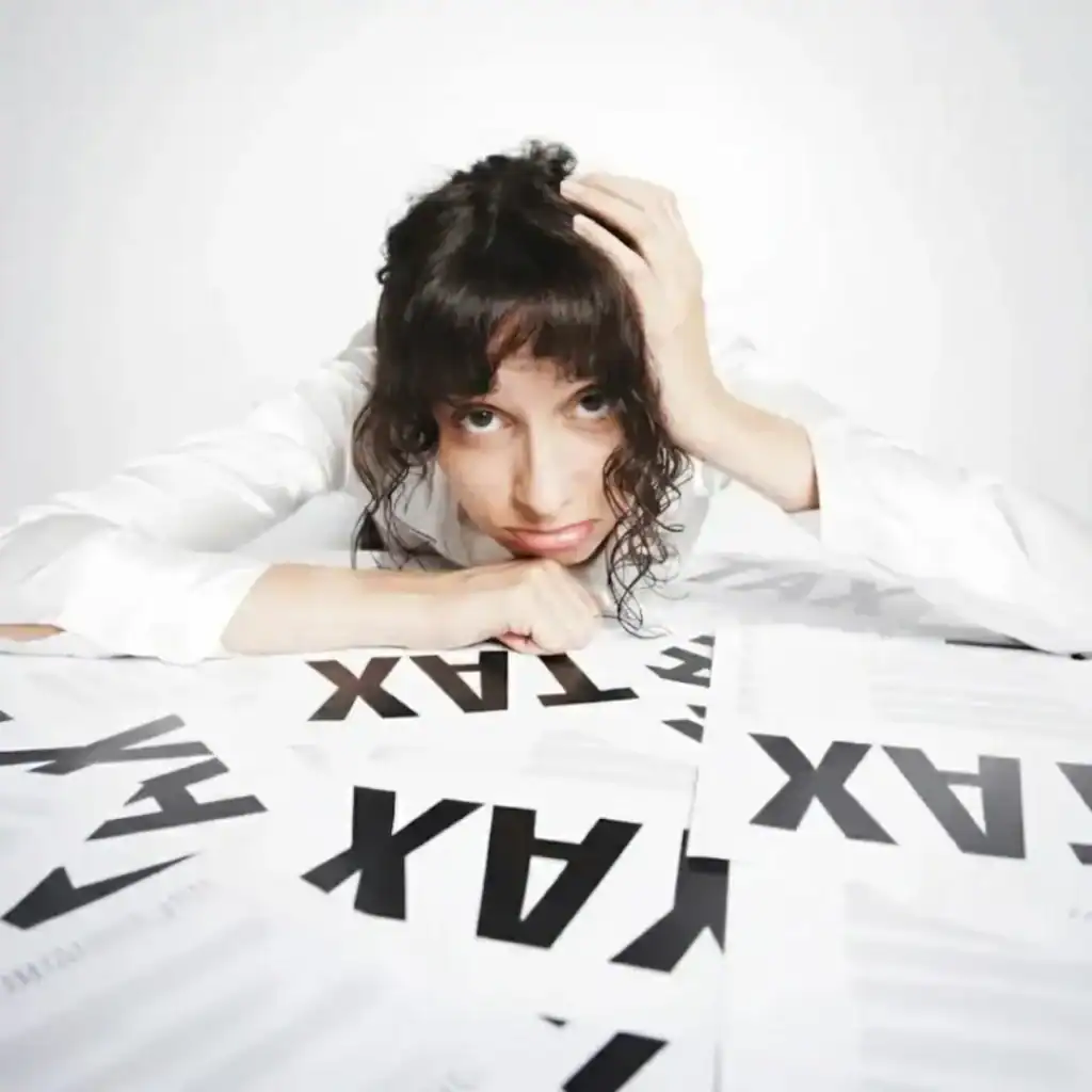 A woman with dark, curly hair rests her head on her hands on a white table, looking distressed or frustrated. She is surrounded by several pieces of paper with the bold, black word "TAX" printed repeatedly across them.